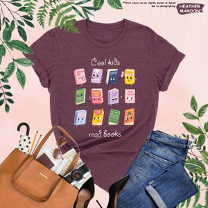 Puede incluir: Camiseta granate con la frase "Cool kids read books." La camiseta presenta un gr&aacute;fico colorido de libros de dibujos animados. Tambi&eacute;n se ve un bolso tote marr&oacute;n, gafas de sol y vaqueros azules.