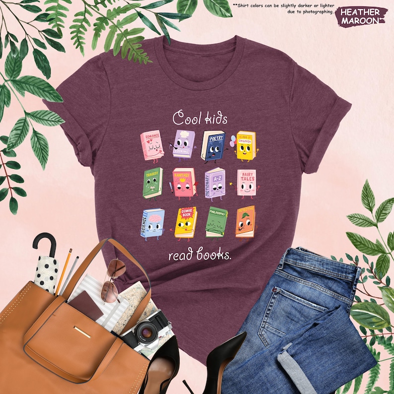 Puede incluir: Camiseta granate con la frase "Cool kids read books." La camiseta presenta un gr&aacute;fico colorido de libros de dibujos animados. Tambi&eacute;n se ve un bolso tote marr&oacute;n, gafas de sol y vaqueros azules.