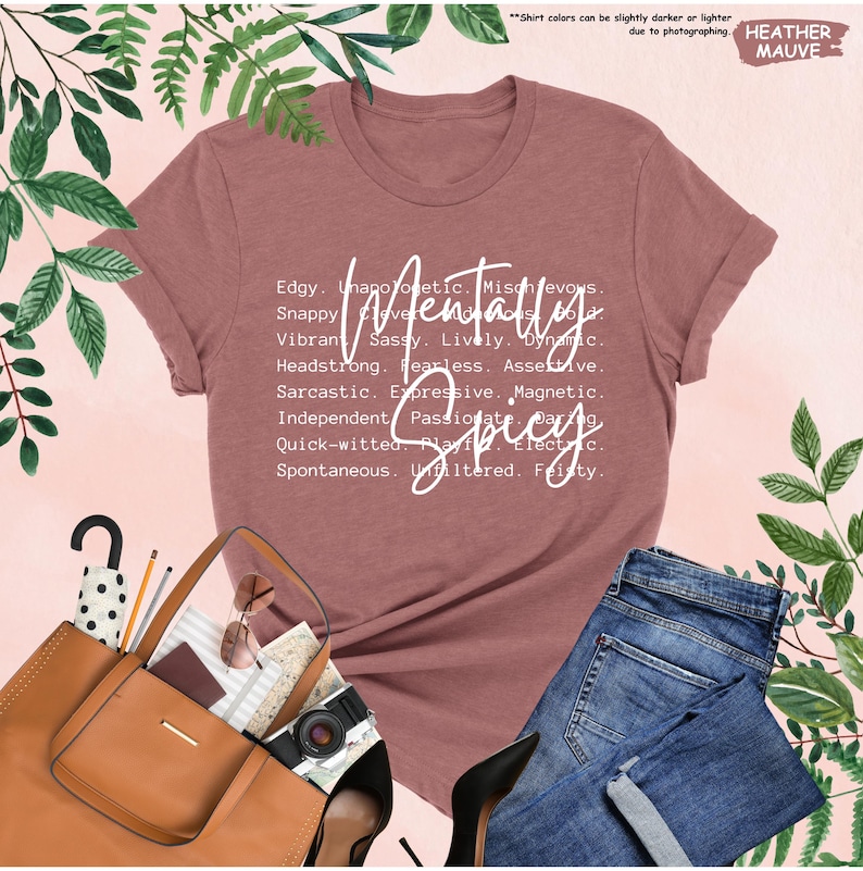 K&ouml;nnte beinhalten: Heather-Mauve-T-Shirt mit dem Schriftzug "Mentally Spicy" in wei&szlig;er Schrift. Das Shirt zeigt eine Liste beschreibender W&ouml;rter. Eine braune Handtasche, Sonnenbrille und Jeans sind im Bild.