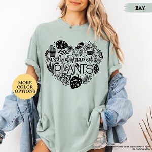 Pu&ograve; includere: Una maglietta verde chiaro con un design a forma di cuore nero che presenta il testo "easily distracted by PLANTS" e varie illustrazioni di piante.