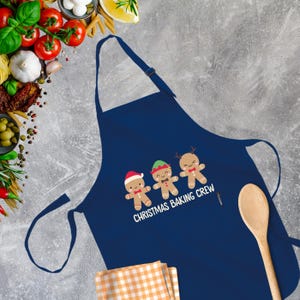 Op de afbeelding: Een marineblauwe schort met een wit ontwerp van drie gingerbreadmen die kerstpetten dragen en de tekst "Christmas Baking Crew".