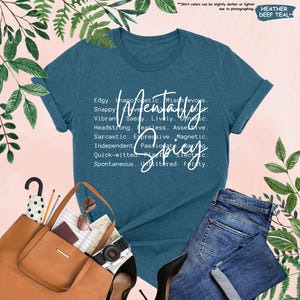 K&ouml;nnte beinhalten: Ein teal-farbenes T-Shirt mit dem Schriftzug "Mentally Spicy" in wei&szlig;er Schrift. Das Shirt zeigt eine Liste von Adjektiven. Das Shirt wird mit einer blauen Jeans, einer braunen Tragetasche und Accessoires kombiniert.