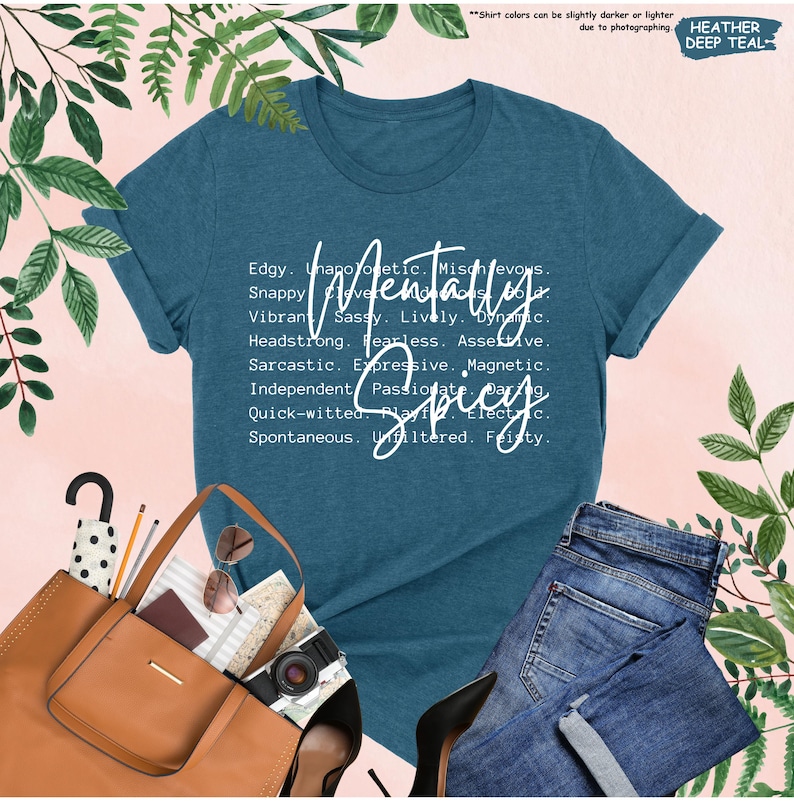 K&ouml;nnte beinhalten: Ein teal-farbenes T-Shirt mit dem Schriftzug "Mentally Spicy" in wei&szlig;er Schrift. Das Shirt zeigt eine Liste von Adjektiven. Das Shirt wird mit einer blauen Jeans, einer braunen Tragetasche und Accessoires kombiniert.