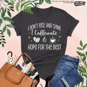 May include: Dark grey heather t-shirt with the text "I DON'T RISE AND SHINE I Caffeinate & HOPE FOR THE BEST" in white. The design includes coffee beans and a coffee cup. The shirt is paired with blue jeans, a brown purse, and black heels.