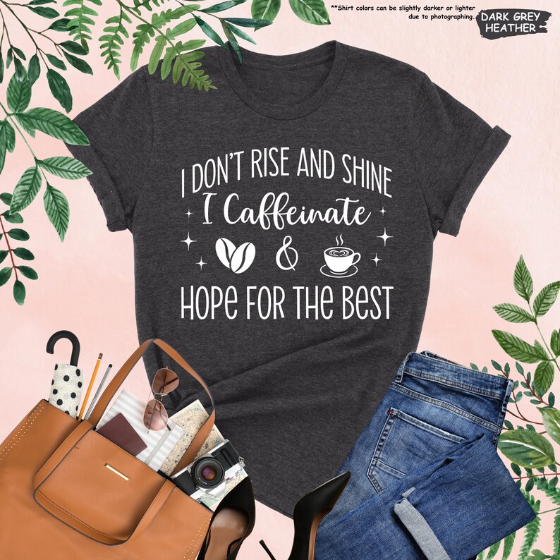 May include: Dark grey heather t-shirt with the text "I DON'T RISE AND SHINE I Caffeinate & HOPE FOR THE BEST" in white. The design includes coffee beans and a coffee cup. The shirt is paired with blue jeans, a brown purse, and black heels.