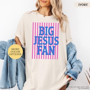 May include: Ivory-colored t-shirt with the text "BIG JESUS FAN" in blue letters, set against a pink striped background. The shirt has rolled-up sleeves and a round neckline. A button with the text "MORE COLOR OPTIONS" is visible.