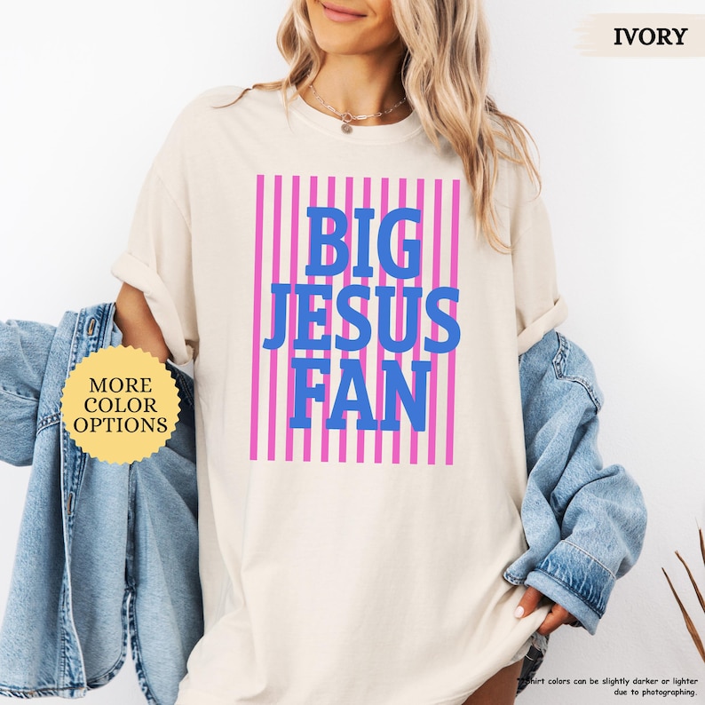 May include: Ivory-colored t-shirt with the text "BIG JESUS FAN" in blue letters, set against a pink striped background. The shirt has rolled-up sleeves and a round neckline. A button with the text "MORE COLOR OPTIONS" is visible.