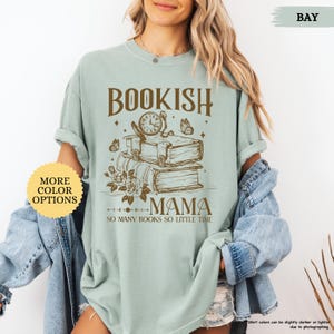 May include: A pale green t-shirt featuring the text "BOOKISH MAMA" above a graphic of books, a clock, and butterflies. The phrase "SO MANY BOOKS SO LITTLE TIME" is below. A button on the left says "MORE COLOR OPTIONS".