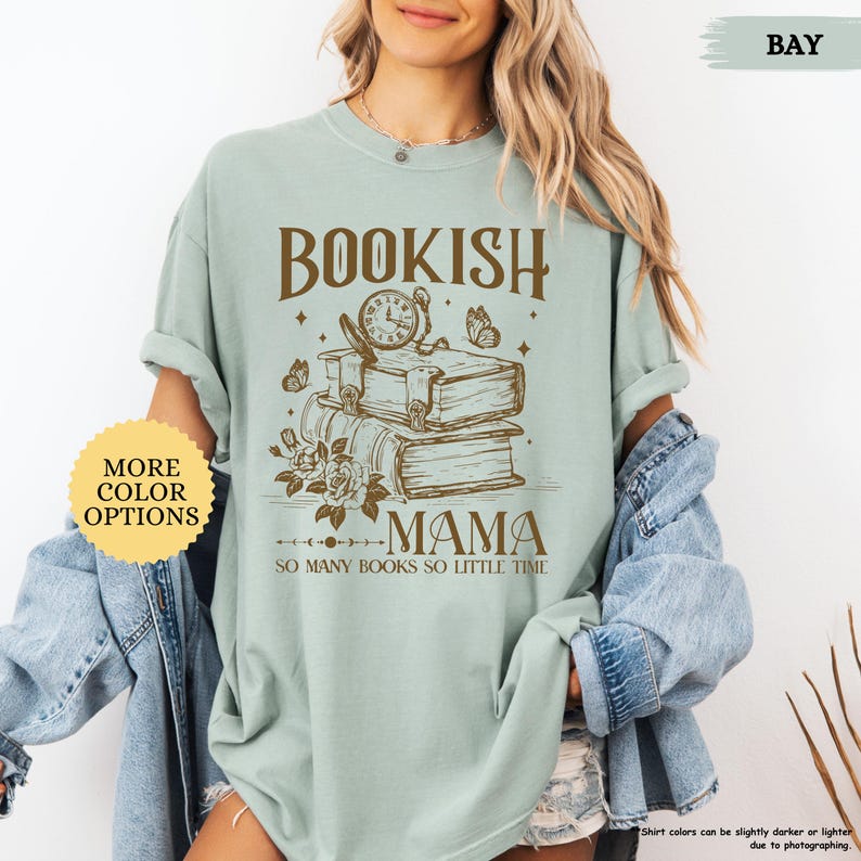May include: A pale green t-shirt featuring the text "BOOKISH MAMA" above a graphic of books, a clock, and butterflies. The phrase "SO MANY BOOKS SO LITTLE TIME" is below. A button on the left says "MORE COLOR OPTIONS".