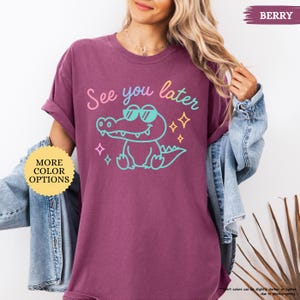 Puede incluir: Camiseta de color baya con la frase "See you later" y un cocodrilo de dibujos animados con gafas de sol. El cocodrilo est&aacute; delineado en turquesa, con texto rosa y amarillo. La camiseta tambi&eacute;n tiene un bot&oacute;n "More Color Options".