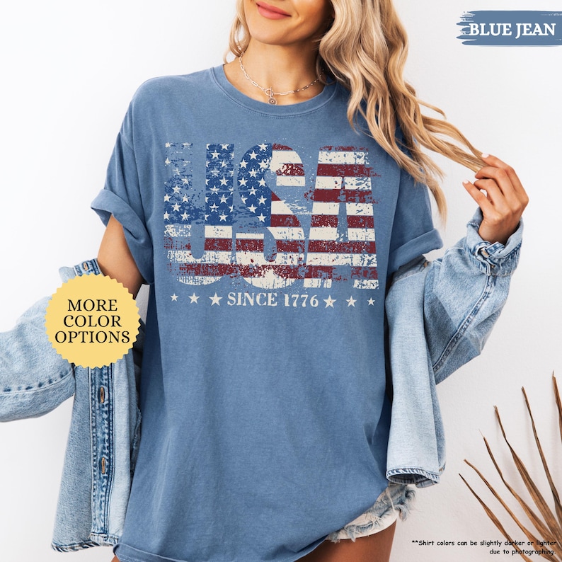 May include: Blue jean-colored t-shirt with a distressed USA flag design. The word "USA" is prominently displayed, with the stars and stripes design. The text "SINCE 1776" is below the flag design. The shirt has rolled-up sleeves.