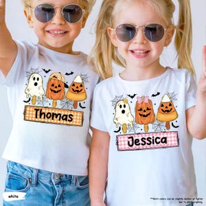 May include: Two white t-shirts featuring Halloween-themed graphics of a ghost, a jack-o'-lantern, and candy corn on sticks. The left shirt displays the name Thomas on a plaid rectangle, while the right shirt displays the name Jessica on a plaid rectangle.