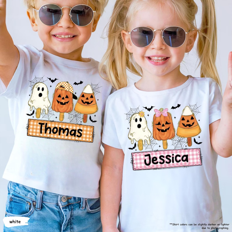 Może przedstawiać: Dwie białe koszulki z grafikami o tematyce Halloween przedstawiającymi ducha, dynię i cukierki kukurydziane na patykach. Na lewej koszulce widnieje imię Thomas na prostokącie w kratkę, a na prawej imię Jessica na prostokącie w kratkę.