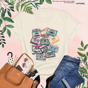 May include: Cream-colored t-shirt featuring a colorful graphic of retro cassette tapes with song titles like "Jesus Anthems" and "Reckless Love." The shirt is paired with blue jeans and a brown tote bag. The text "Natural" is in the upper right corner.