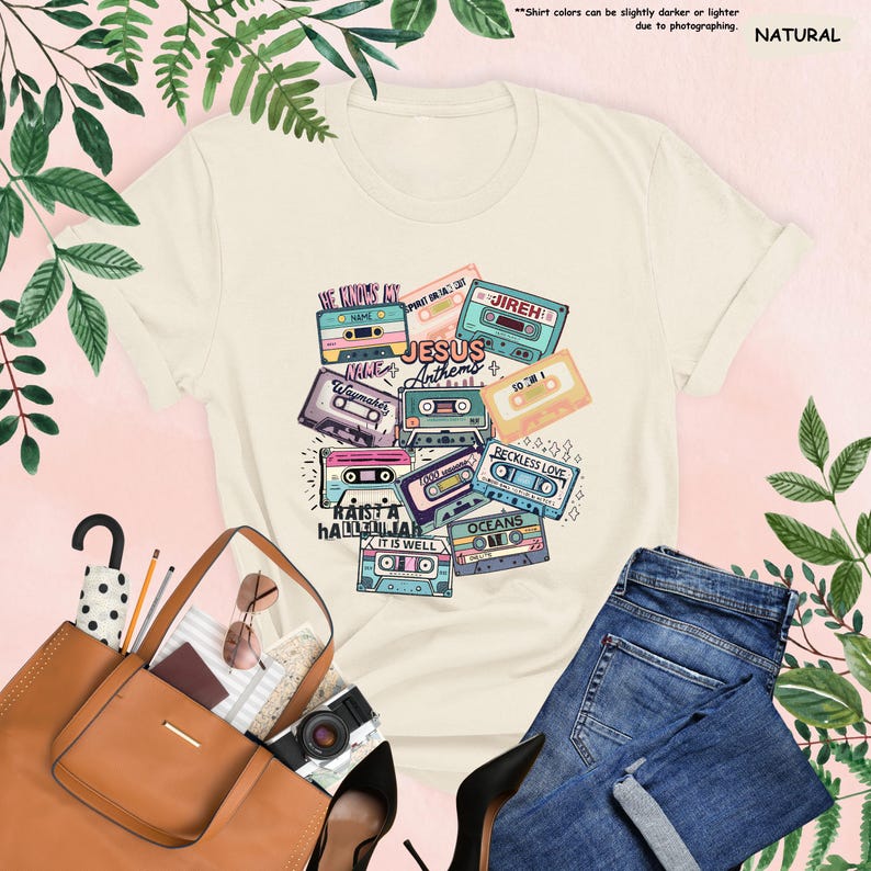 May include: Cream-colored t-shirt featuring a colorful graphic of retro cassette tapes with song titles like "Jesus Anthems" and "Reckless Love." The shirt is paired with blue jeans and a brown tote bag. The text "Natural" is in the upper right corner.