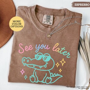 Puede incluir: Camiseta color espresso con un cocodrilo de dibujos animados con gafas de sol y la frase "See you later" en colores pastel. La camiseta tiene cuello redondo y mangas cortas. Un collar plateado con un colgante est&aacute; sobre la camiseta.