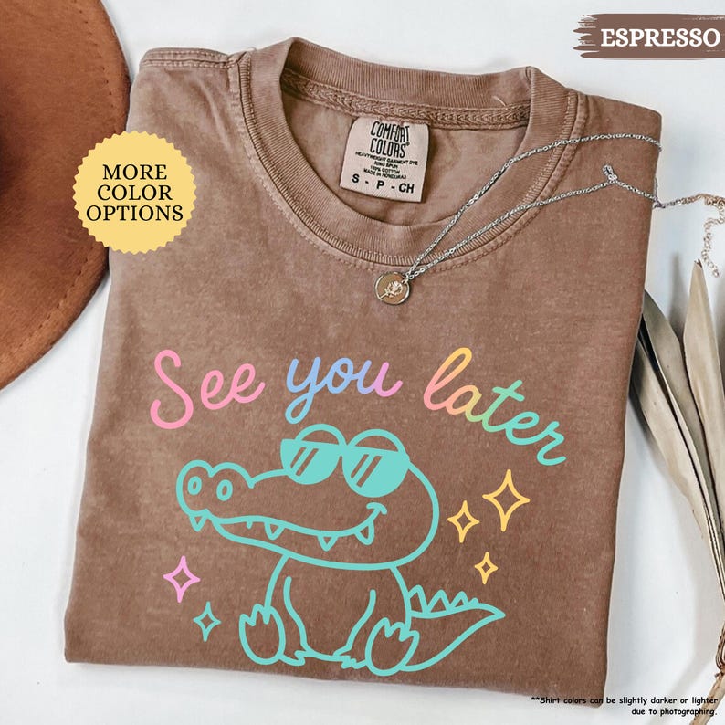 Puede incluir: Camiseta color espresso con un cocodrilo de dibujos animados con gafas de sol y la frase "See you later" en colores pastel. La camiseta tiene cuello redondo y mangas cortas. Un collar plateado con un colgante est&aacute; sobre la camiseta.