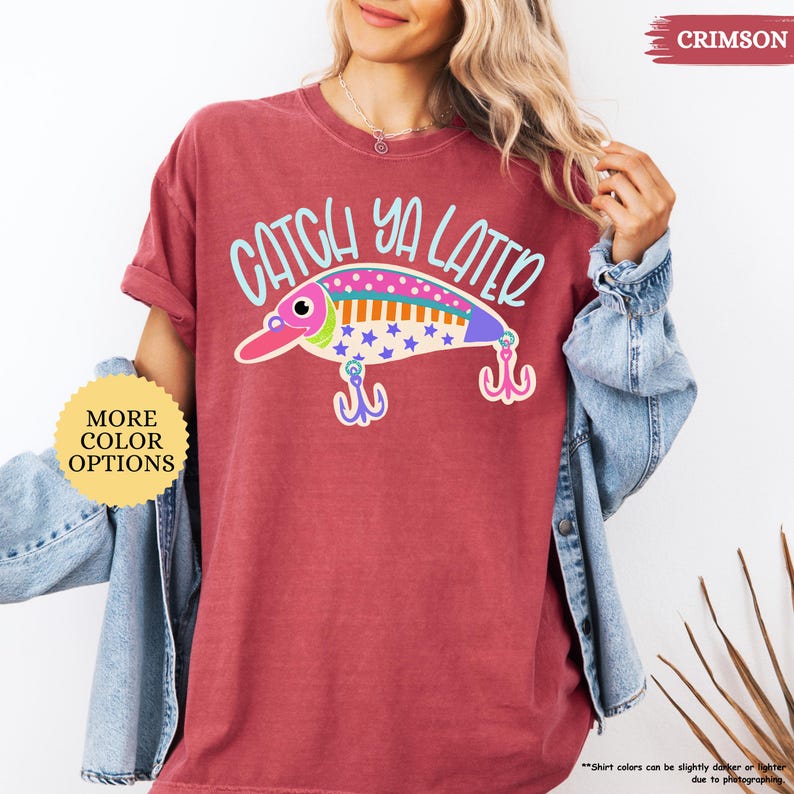 以下が含まれることがあります： クリムゾンカラーの半袖Tシャツ。水色で「CATCH YA LATER」という文字と、カラフルな釣り用ルアーのグラフィックが描かれています。ルアーのデザインにはピンク、オレンジ、紫のアクセントがあり、クリーム色の背景に青い星の模様があしらわれています。