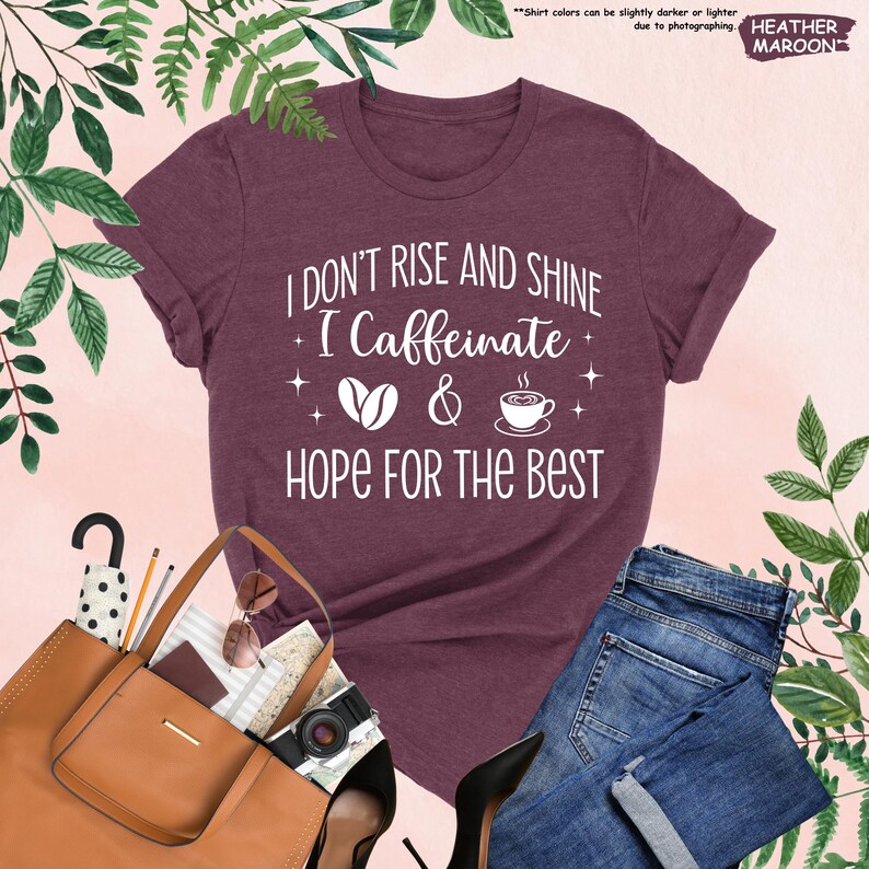 May include: Heather maroon t-shirt with the text "I don't rise and shine, I caffeinate & hope for the best" in white. The design includes coffee beans and a coffee cup. The shirt is paired with blue jeans and a brown handbag.