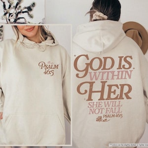 Puede incluir: Sudadera con capucha color arena con bolsillo delantero y capucha con cord&oacute;n. La parte delantera presenta el texto "Psalm 46:5" en escritura marr&oacute;n. La parte trasera muestra "God is Within Her" y "She Will Not Fall" en letras marrones y rosas, con "Psalm 46:5" debajo.