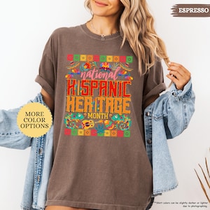 Peut inclure: T-shirt marron avec l'inscription "national HISPANIC HERITAGE MONTH" dans un design color&eacute;. Le t-shirt a un col rond et des manches courtes. Un bouton sur le c&ocirc;t&eacute; gauche indique "MORE COLOR OPTIONS".