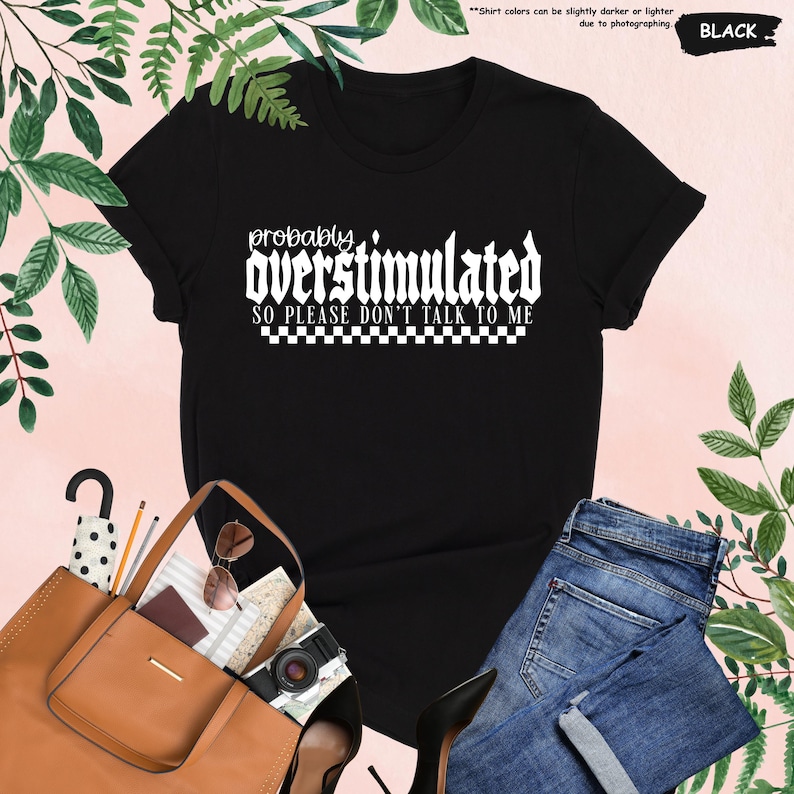 May include: Black t-shirt with white text that reads "probably overstimulated so please don't talk to me". The text is in a gothic font and is surrounded by a checkered pattern.