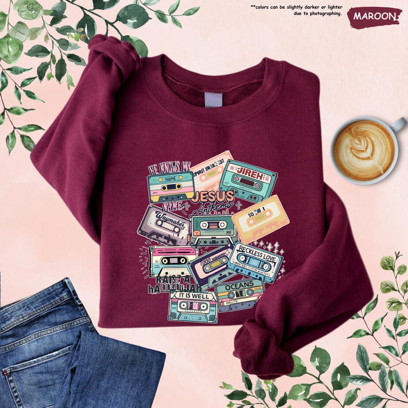 May include: Maroon sweatshirt with a graphic of cassette tapes. The tapes have phrases like "Jesus", "Reckless Love", and "Oceans". The sweatshirt is folded with a pair of blue jeans and a cup of coffee.