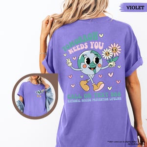 May include: A violet t-shirt featuring a cartoon Earth character with flowers, the text reads "TOMORROW NEEDS YOU" and "CALL OR TEXT 988 NATIONAL SUICIDE PREVENTION LIFELINE". The shirt is a solid violet colour.