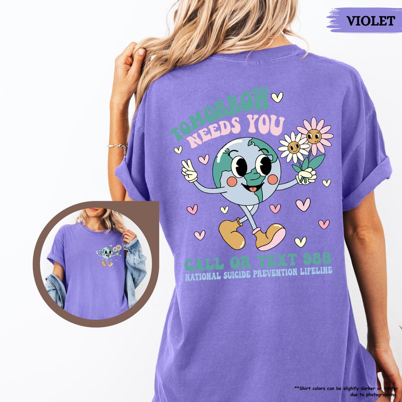 May include: A violet t-shirt featuring a cartoon Earth character with flowers, the text reads "TOMORROW NEEDS YOU" and "CALL OR TEXT 988 NATIONAL SUICIDE PREVENTION LIFELINE". The shirt is a solid violet colour.