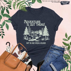 May include: Heather navy t-shirt with the text "ADVENTURE IS OUT THERE! BUT SO ARE SERIAL KILLERS" and a graphic of a forest scene. The shirt is paired with blue jeans and a brown tote bag.