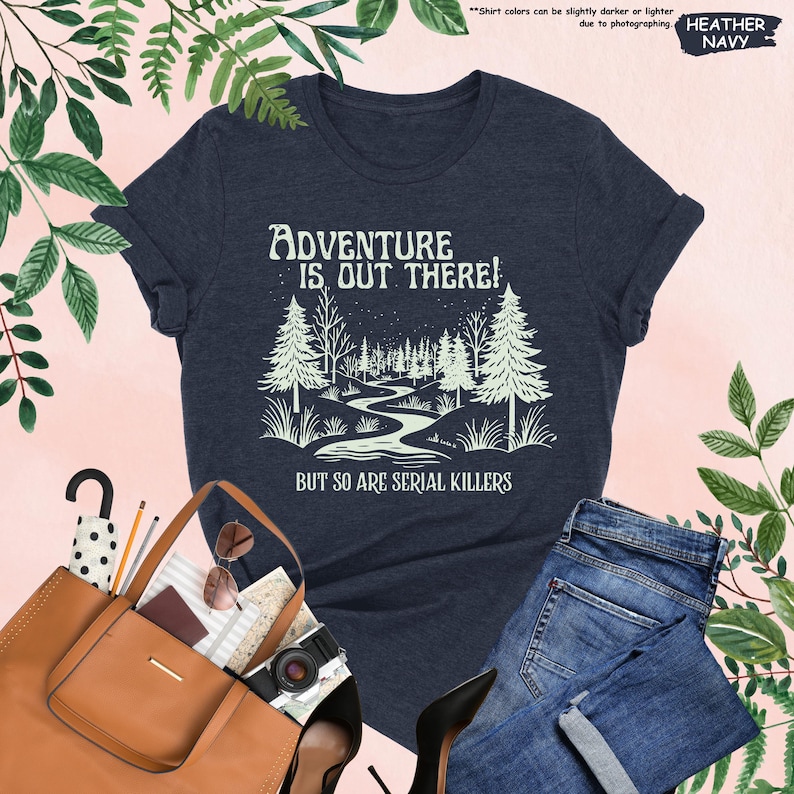May include: Heather navy t-shirt with the text "ADVENTURE IS OUT THERE! BUT SO ARE SERIAL KILLERS" and a graphic of a forest scene. The shirt is paired with blue jeans and a brown tote bag.