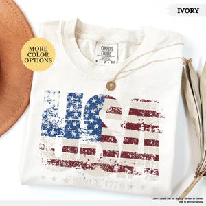 May include: Ivory-colored t-shirt with a distressed USA flag design. The shirt features the text "USA" in a patriotic red, white, and blue color scheme, with "SINCE 1776" printed below. A gold necklace and a brown hat are also visible.