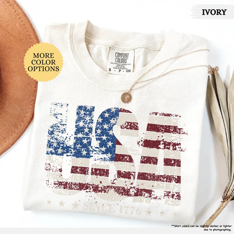 May include: Ivory-colored t-shirt with a distressed USA flag design. The shirt features the text "USA" in a patriotic red, white, and blue color scheme, with "SINCE 1776" printed below. A gold necklace and a brown hat are also visible.