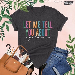 May include: A dark heather grey t-shirt with the text "LET ME TELL YOU ABOUT my Jesus" in rainbow colors. The text is stylized with floral accents.