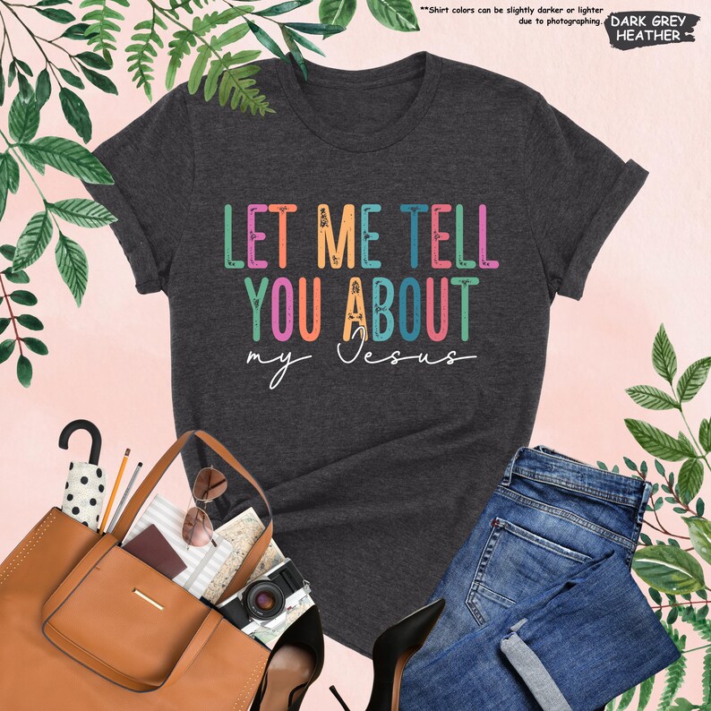 May include: A dark heather grey t-shirt with the text "LET ME TELL YOU ABOUT my Jesus" in rainbow colors. The text is stylized with floral accents.