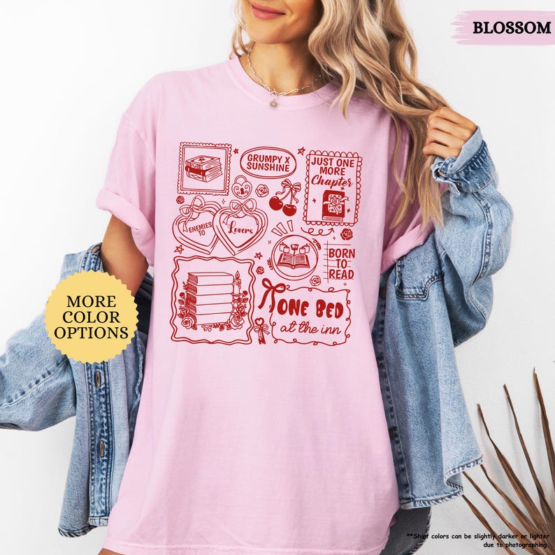 Puede incluir: Una camiseta rosa claro con un dise&ntilde;o gr&aacute;fico rojo que presenta ilustraciones y texto con tem&aacute;tica de libros. El dise&ntilde;o incluye frases como "One Bed at the Inn" y "Just One More Chapter". La camiseta tambi&eacute;n tiene un bot&oacute;n de "More Color Options".