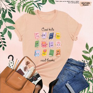 Puede incluir: Una camiseta color melocot&oacute;n con el texto "Cool kids read books." La camiseta presenta un gr&aacute;fico colorido de libros animados. La camiseta se combina con vaqueros azules, un bolso marr&oacute;n y tacones negros.