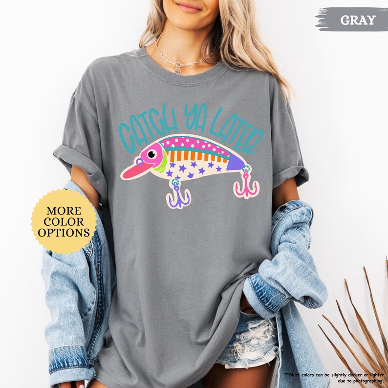 以下が含まれることがあります： グレーの半袖Tシャツ。カラフルな釣り用ルアーのグラフィックと、ティールカラーで「CATCH YA LATER」の文字がプリントされています。ルアーのデザインには、星やドット柄のピンク、オレンジ、ブルーの模様があり、2つのピンクの釣り針が描かれています。