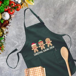Op de afbeelding: Groene kerstbakschort met een zak en drie gingerbreadmen met verschillende hoeden. De tekst op het schort luidt "Christmas Baking Crew".