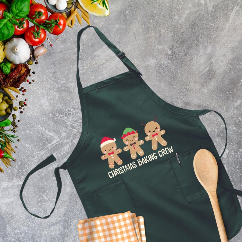 Op de afbeelding: Groene kerstbakschort met een zak en drie gingerbreadmen met verschillende hoeden. De tekst op het schort luidt "Christmas Baking Crew".