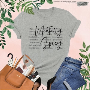 K&ouml;nnte beinhalten: Graues Athletic Heather T-Shirt mit dem Schriftzug "Mentally Spicy" in schwarzer Schrift. Das Shirt listet auch Adjektive wie "Edgy", "Sassy" und "Fearless" auf. Das Bild zeigt auch eine blaue Jeans, eine braune Handtasche und schwarze High Heels.