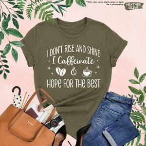 May include: Olive green t-shirt with the text "I DON'T RISE AND SHINE I Caffeinate & HOPE FOR THE BEST" in white. The shirt is paired with blue jeans, a brown handbag, and black heels. The image includes a camera, sunglasses, and an umbrella.