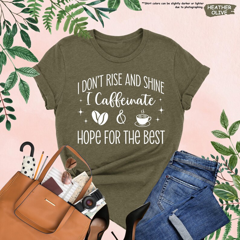 May include: Olive green t-shirt with the text "I DON'T RISE AND SHINE I Caffeinate & HOPE FOR THE BEST" in white. The shirt is paired with blue jeans, a brown handbag, and black heels. The image includes a camera, sunglasses, and an umbrella.