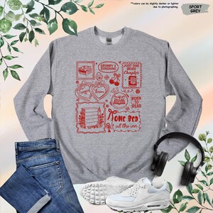 May include: Sport grey sweatshirt with a red graphic design featuring book-themed illustrations and text. The text includes phrases like "Grumpy x Sunshine," "Just One More Chapter," and "Born to Read."