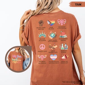 May include: A terracotta-colored t-shirt with various graphic designs and text. The back of the shirt features icons and phrases such as "Black Lives Matter," "Love is Love," and "Science is Real." The front of the shirt in the inset image says "big fan of human rights."