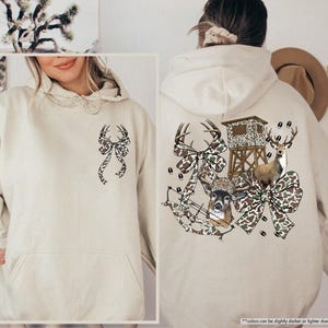 May include: A sand-colored hoodie with a front pocket and a drawstring hood. The front features a bow design with deer antlers. The back of the hoodie has a hunting-themed graphic with deer, bows, and a hunting blind.