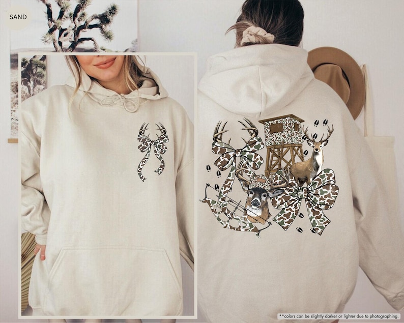 May include: A sand-colored hoodie with a front pocket and a drawstring hood. The front features a bow design with deer antlers. The back of the hoodie has a hunting-themed graphic with deer, bows, and a hunting blind.