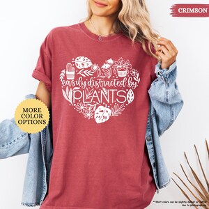 Pu&ograve; includere: Una maglietta color cremisi con un design a forma di cuore bianco che presenta piante e il testo "easily distracted by PLANTS".