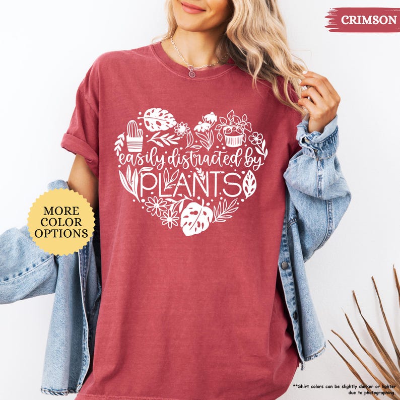 Pu&ograve; includere: Una maglietta color cremisi con un design a forma di cuore bianco che presenta piante e il testo "easily distracted by PLANTS".