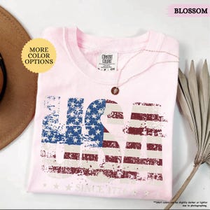 May include: A light pink t-shirt with a distressed USA graphic in blue, white, and red. The text "SINCE 1776" is below the graphic. The shirt is folded and has a Comfort Colors tag. A gold necklace with a pendant is on the shirt.